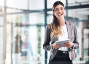 Eine Frau in Business-Kleidung steht in einem modernen Büro, lächelt und hält ein digitales Tablet. Der Hintergrund ist hell mit Glaswänden und unscharfen Figuren, die eine professionelle Atmosphäre vermitteln.