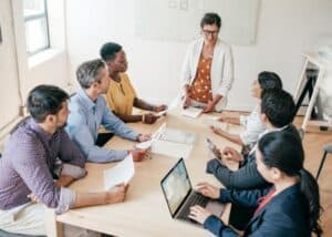 Eine Gruppe von sieben Personen sitzt um einen Tisch in einem hellen Büro und führt eine Besprechung durch. Eine Frau steht am Kopfende des Tisches, lächelt und hält Papiere in der Hand, während die anderen zuhören und Notizen machen.