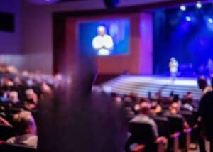 Ein unscharfes Bild zeigt ein Publikum, das in einem großen Auditorium sitzt und auf eine Bühne blickt, auf der eine Person unter blauer und violetter Beleuchtung steht, während im Hintergrund ein großer Bildschirm eine weitere Person anzeigt.