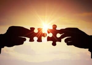 Zwei silhouettierte Hände halten einander passende Puzzleteile vor einem leuchtenden Sonnenuntergangshimmel entgegen, wobei die Sonne durch die Lücke scheint, in der sich die Teile treffen.