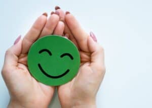 Ein Paar Hände, die sanft einen grünen Papierkreis schröpfen, auf dem ein lächelndes Gesicht gezeichnet ist, das Glück oder Positivität symbolisiert, vor einem schlichten weißen Hintergrund.