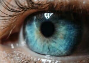 Nahaufnahme eines blauen menschlichen Auges mit detaillierten Irismustern, sichtbaren Wimpern und Lichtreflexen auf der Augenoberfläche.