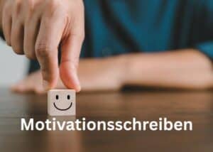 Eine Hand legt einen Holzblock mit einem Smiley-Gesicht auf einen Tisch. Das Wort Motivationsschreiben erscheint in fetter wei&szlig;er Schrift am unteren Rand des Bildes.