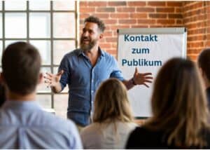 Ein Mann steht vor einer Gruppe, spricht und gestikuliert mit einem L&auml;cheln. Hinter ihm steht ein Flipchart mit der Aufschrift "Kontakt zum Publikum".