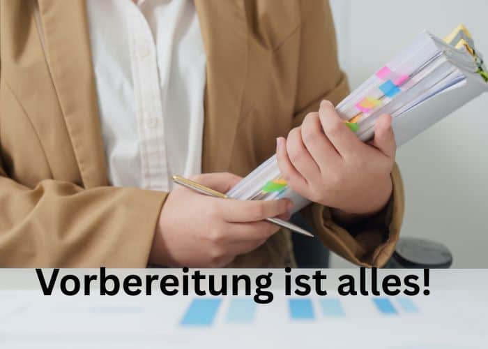 Eine Person in einem braunen Blazer, die eine Mappe mit bunten Registerstreifen in der Hand hält; über dem Bild steht auf Deutsch „Vorbereitung ist alles!“, was auf Englisch „Preparation is everything!“ bedeutet.