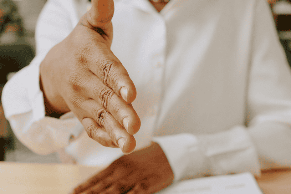 Eine Person in einem weißen Hemd streckt über einen Tisch hinweg, auf dem ein Lebenslauf und ein Foto liegen, die Hand zum Handschlag aus. Der Text darüber lautet „Ein überzeugender Lebenslauf“ auf gelbem Hintergrund.