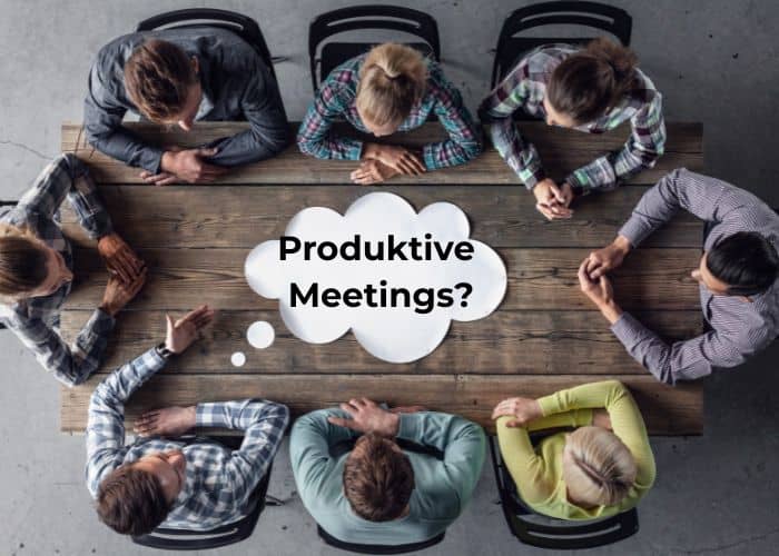 Sieben Personen sitzen um einen Holztisch herum (Ausblick von oben), auf dem eine Sprechblase mit der Aufschrift „Produktive Meetings?“ in schwarzer Schrift zu sehen ist.
