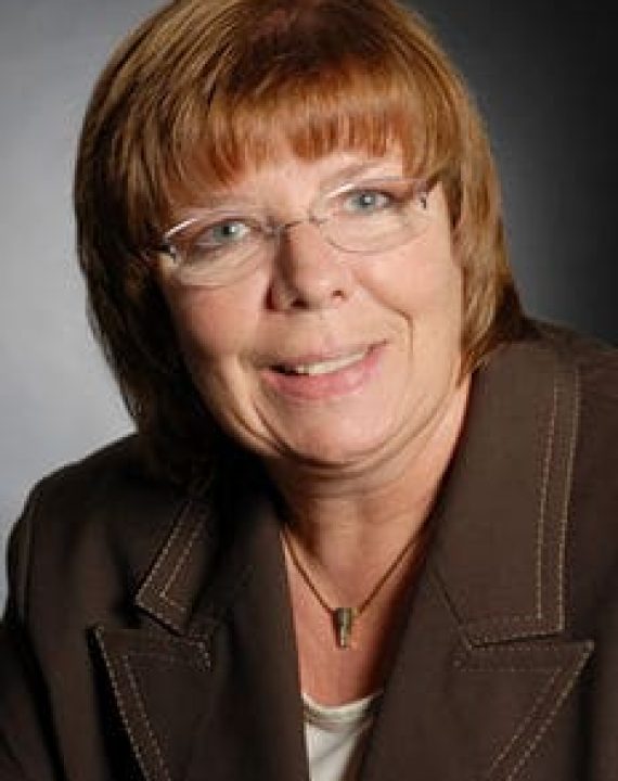 Eine Frau mit kurzen, hellbraunen Haaren und Brille l&auml;chelt. Sie tr&auml;gt einen braunen Blazer &uuml;ber einem wei&szlig;en Oberteil, mit einem dunklen Hintergrund hinter ihr - perfekt, um Jobcoaching M&uuml;nchen zu repr&auml;sentieren.