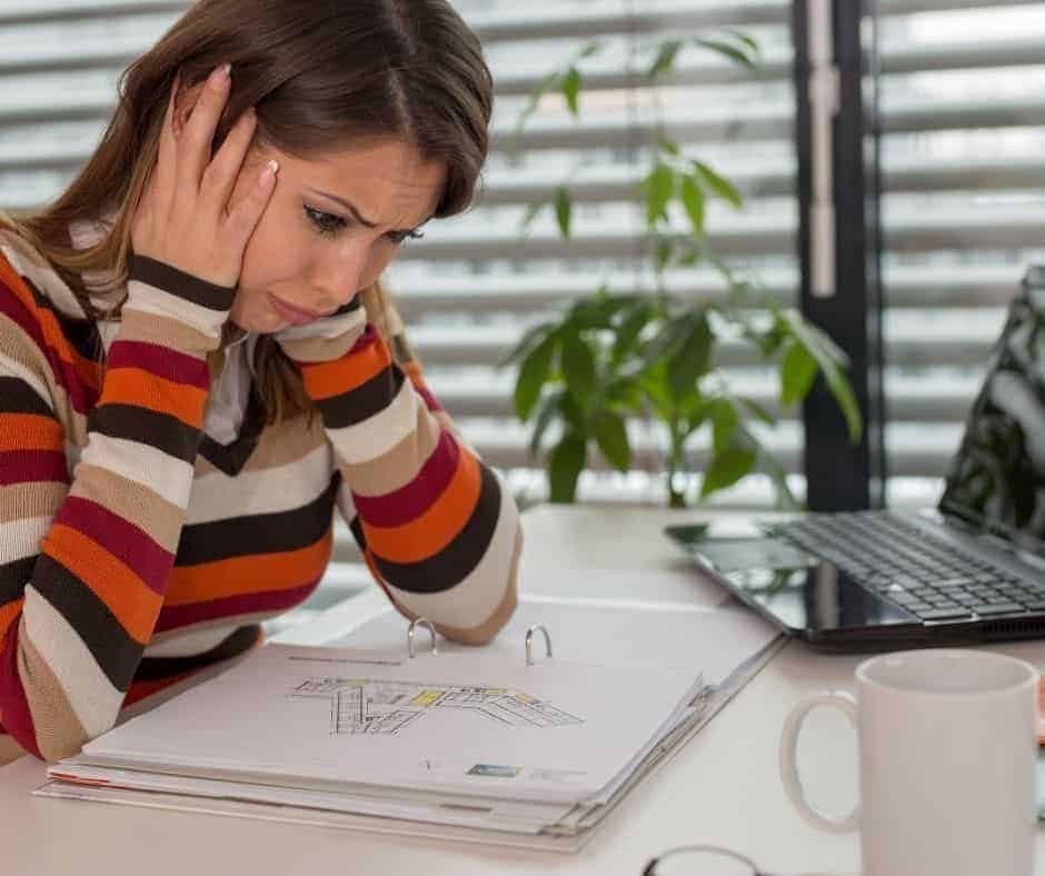 Eine Frau in einem bunt gestreiften Pullover sitzt in M&uuml;nchen an ihrem Schreibtisch, h&auml;lt sich gestresst den Kopf und starrt auf Papiere und einen Laptop - vielleicht erw&auml;gt sie ein Jobcoaching M&uuml;nchen, um ihr Arbeitspensum zu bew&auml;ltigen. In der N&auml;he stehen ein Kaffeebecher und eine Pflanze.
