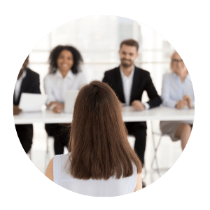 Eine Frau mit langen braunen Haaren sitzt vor einer Runde von vier Personen in Business-Kleidung, was auf ein Vorstellungsgespr&auml;ch oder ein berufliches Treffen hindeutet - ein ideales Szenario f&uuml;r diejenigen, die das Jobcoaching M&uuml;nchen suchen, um ihre Karriereaussichten zu verbessern.
