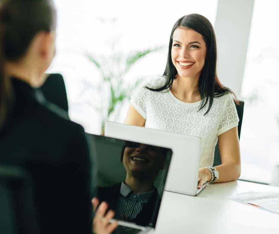 Zwei Frauen sitzen sich an einem Schreibtisch mit Laptop gegen&uuml;ber und f&uuml;hren ein Gespr&auml;ch. Die eine Frau, die in die Kamera schaut, l&auml;chelt freundlich - und f&auml;ngt damit den unterst&uuml;tzenden Geist von Jobcoaching M&uuml;nchen in einer modernen B&uuml;roumgebung ein.
